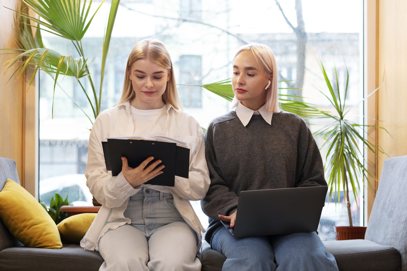Giovani ragazze bionde, vestite casual, sedute di spalle ad una luminosa finestra, una di fianco all'altra, una con in mano un libro, tenuto dietro da un portablocco nero; l'altra con il pc sulle cosce. Quest'ultima osserva le pagine del libro della vicina