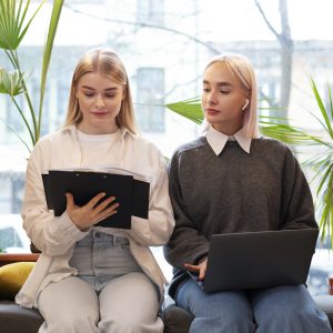 Giovani ragazze bionde, vestite casual, sedute di spalle ad una luminosa finestra, una di fianco all'altra, una con in mano un libro, tenuto dietro da un portablocco nero; l'altra con il pc sulle cosce. Quest'ultima osserva le pagine del libro della vicina